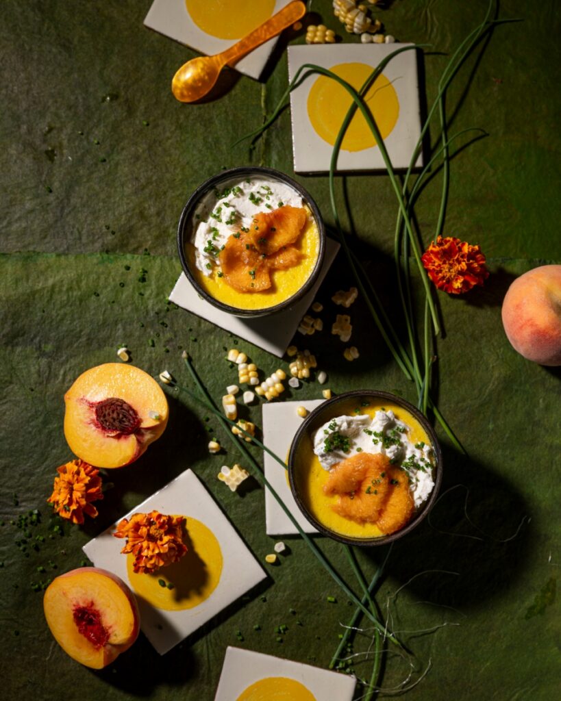 Two bowls of corn custard topped with pickled peaches and whipped goat cheese on a green textured background with fresh peaches and corn kernels.