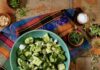 Crushed Cucumber and Avocado Salad A light green bowl with a crushed cucumber and avocado salad inside with pieces of feta cheese and it sits on a pattern cloth on a table.
