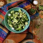 A light green bowl with a crushed cucumber and avocado salad inside with pieces of feta cheese and it sits on a pattern cloth on a table.