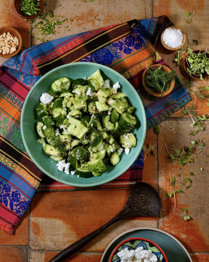 A light green bowl with a crushed cucumber and avocado salad inside with pieces of feta cheese and it sits on a pattern cloth on a table.