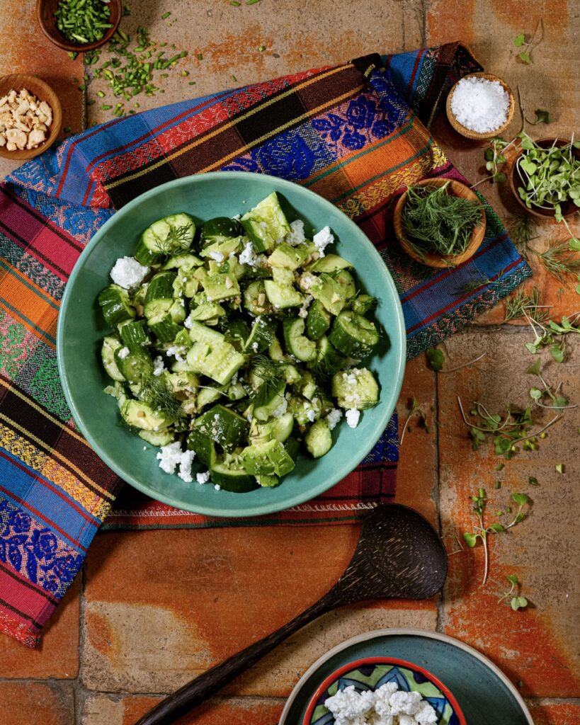 A light green bowl with a crushed cucumber and avocado salad inside with pieces of feta cheese and it sits on a pattern cloth on a table.
