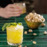 A short cocktail glass with a yellow cocktail and basil garnish next tot a bowl of nuts and a person holding a cocktail behind it.