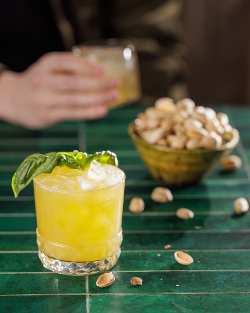 A short cocktail glass with a yellow cocktail and basil garnish next tot a bowl of nuts and a person holding a cocktail behind it.
