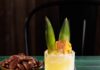 6 Refreshing Summer Cocktail Recipes from New Mexico Mixologists A yellow cocktail in a tall glass with two pineapple leaf garnishes and a bowl of almonds sitting behind the cocktail.