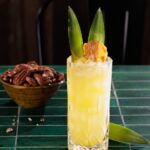 A yellow cocktail in a tall glass with two pineapple leaf garnishes and a bowl of almonds sitting behind the cocktail.