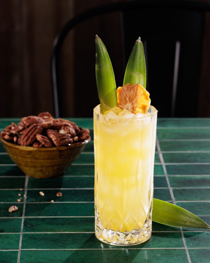 A yellow cocktail in a tall glass with two pineapple leaf garnishes and a bowl of almonds sitting behind the cocktail.
