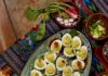 Deviled Eggs An oval plate full of deviled egg halves on top of a pattern cloth surrounded by various toppings for the eggs.