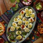 An oval plate full of deviled egg halves on top of a pattern cloth surrounded by various toppings for the eggs.