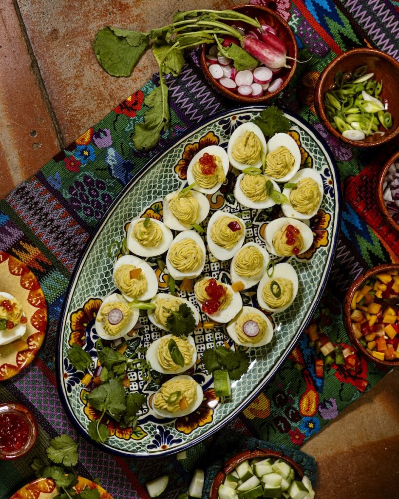 An oval plate full of deviled egg halves on top of a pattern cloth surrounded by various toppings for the eggs.