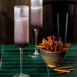 Two tall cocktail flutes with a High Desert Spritz inside with a purple color and a bowl of pretzel sticks beside the cocktails.