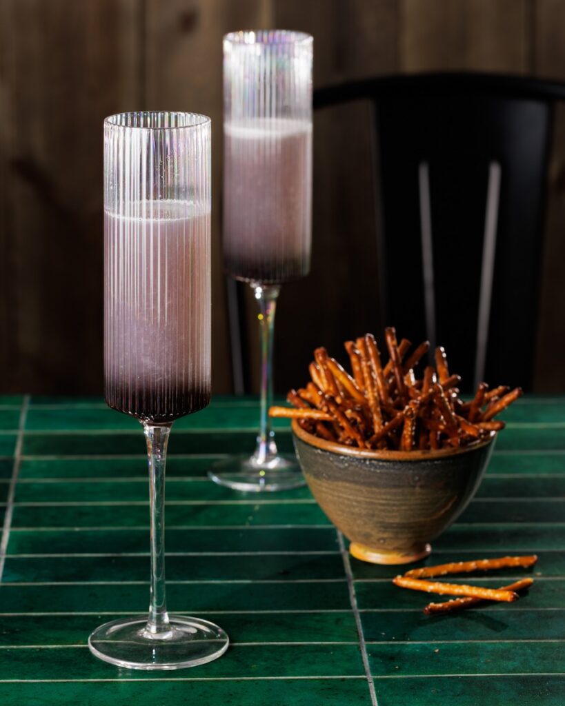 Two tall cocktail flutes with a High Desert Spritz inside with a purple color and a bowl of pretzel sticks beside the cocktails.