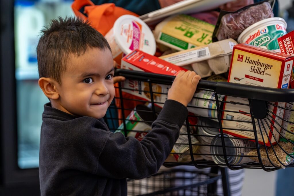 A little boy hangs on to a shopping cart full of food.