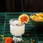 A short cocktail glass with lavender herbs on the rim and a grapefruit slice on the rim too. An orange flower sits beside the glass.