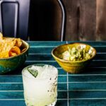 A short cocktail glass with makrut lime leaves as a garnish in front of two bowls of chips and guac.