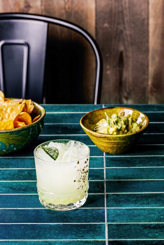 A short cocktail glass with makrut lime leaves as a garnish in front of two bowls of chips and guac.