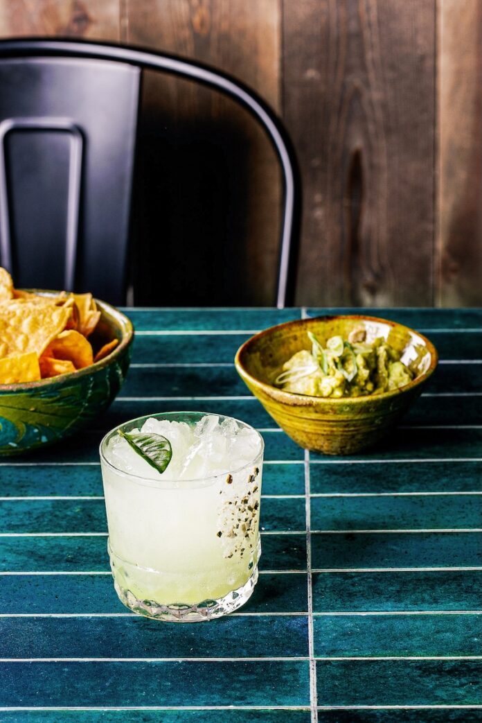Makrut’s Daisy Cocktail With Makrut Lime Leaves TABLE Magazine A short cocktail glass with makrut lime leaves as a garnish in front of two bowls of chips and guac.