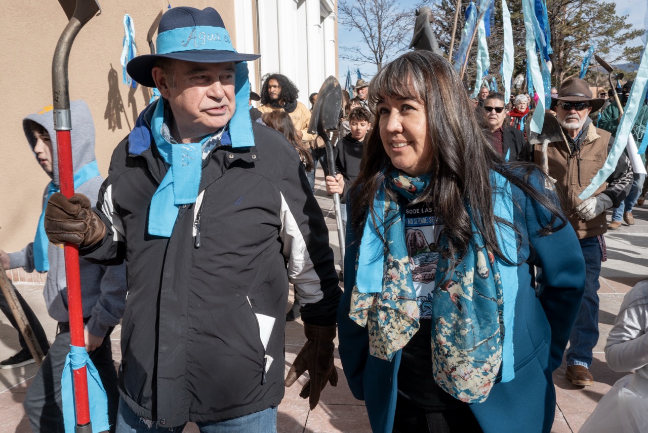 A woman in a blue scarf walks with a man in a blue scarf and hat with a blue ribbon.