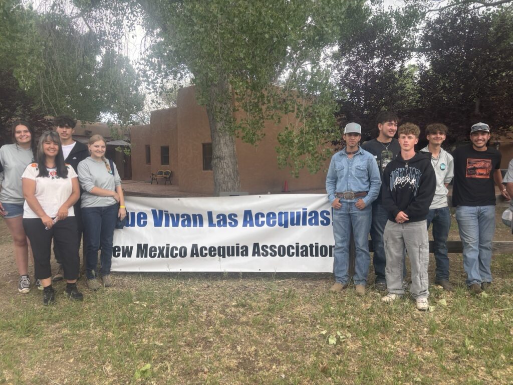 A group of teenagers stands on both sides of a sign for the New Mexico Acequia Association.