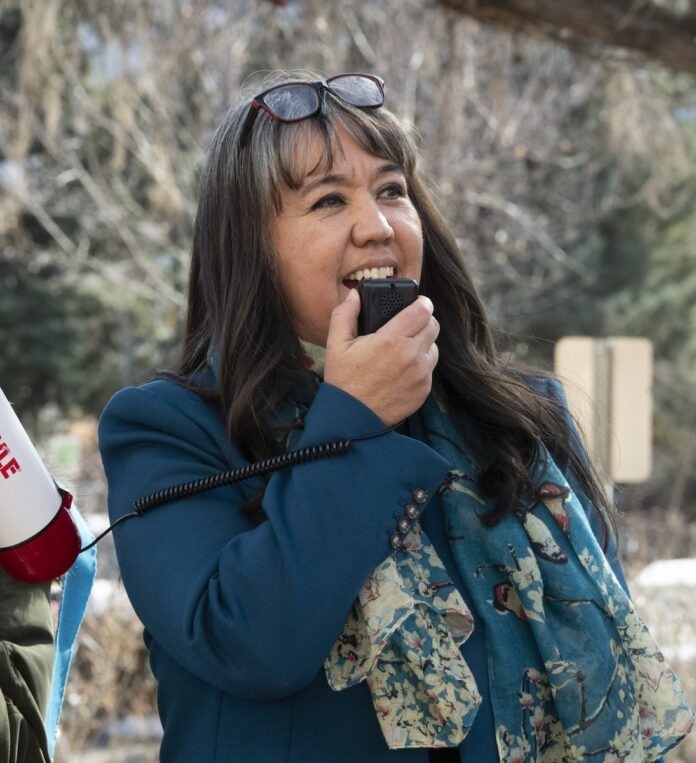 A woman in a teal coat speaks into a megaphone microphone with glasses on top of her head.
