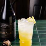 A yellow and clear cocktail with a lemon twist garnish sitting beside a bottle of VARA brut sparkling wine.