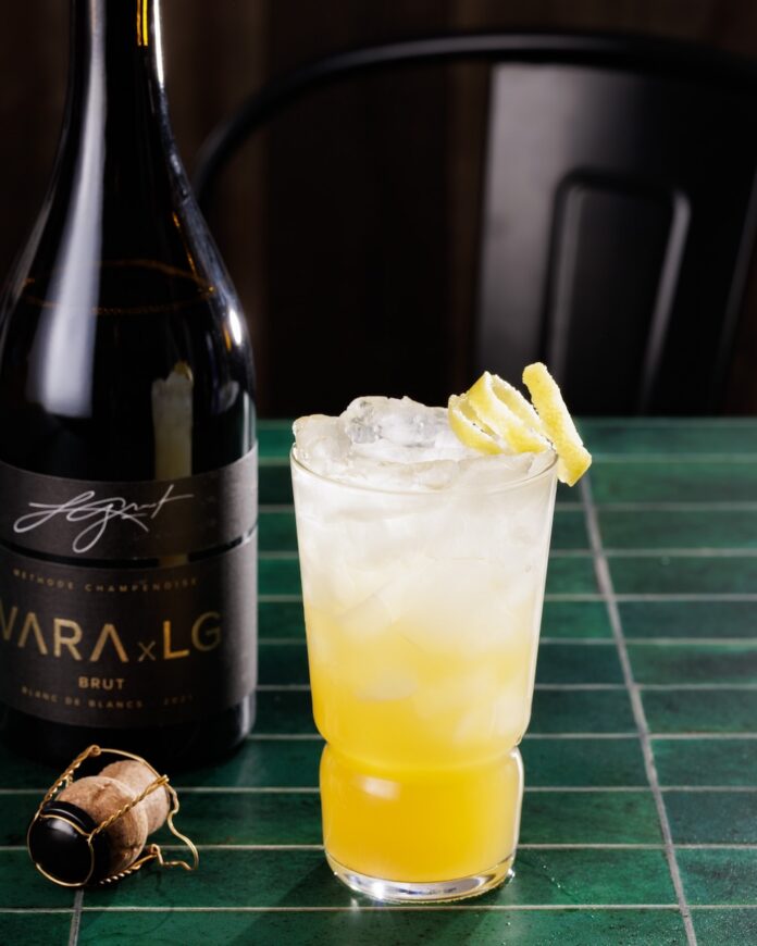 New Mexico Sunrise Brandy and Sparkling Wine TABLE Magazine A yellow and clear cocktail with a lemon twist garnish sitting beside a bottle of VARA brut sparkling wine.