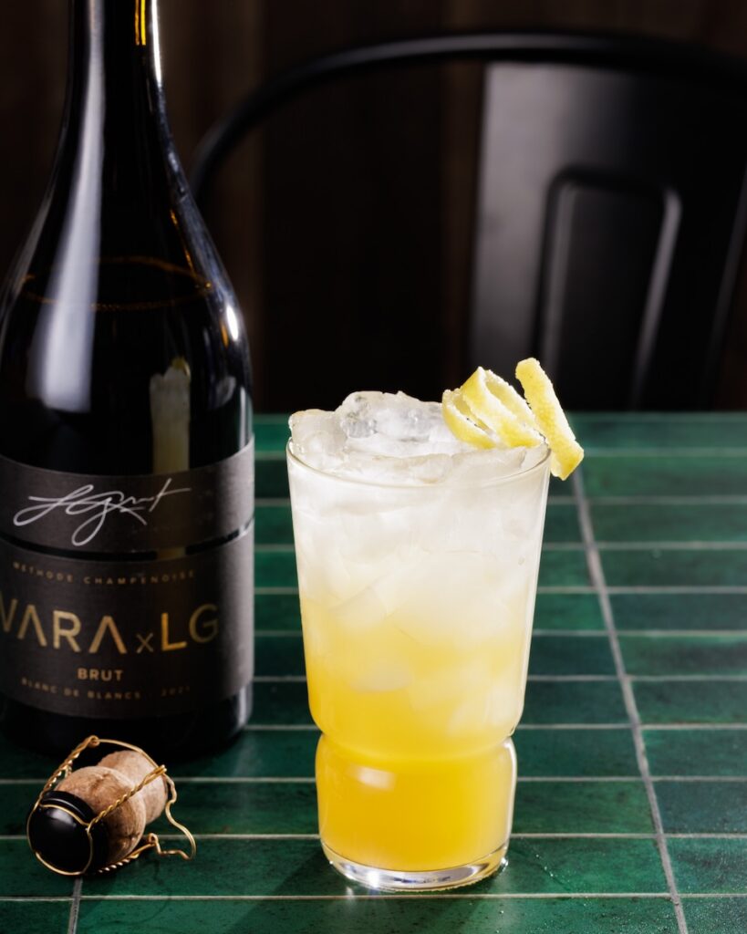 A yellow and clear cocktail with a lemon twist garnish sitting beside a bottle of VARA brut sparkling wine.