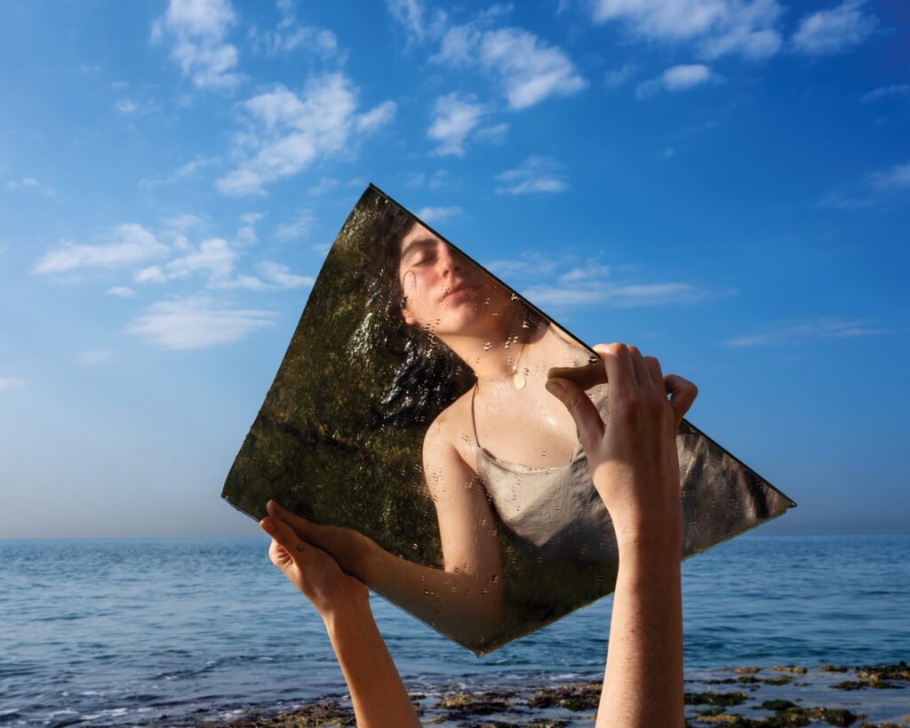 A woman holds a mirror up to her reflection with a blue sky and ocean behind her.