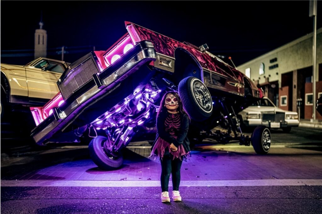 A little girl stands in front of a muscle car held up with lights underneath.