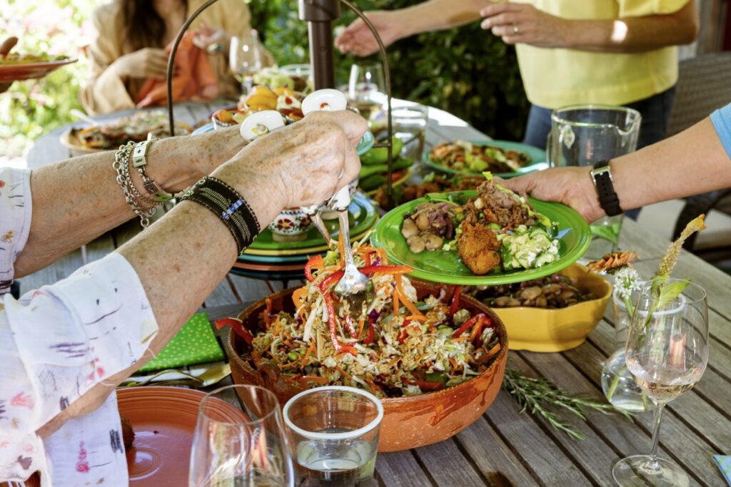 Various plates of potluck foods on a picnic table as a woman mixes a salad and others dive into the other bowls.