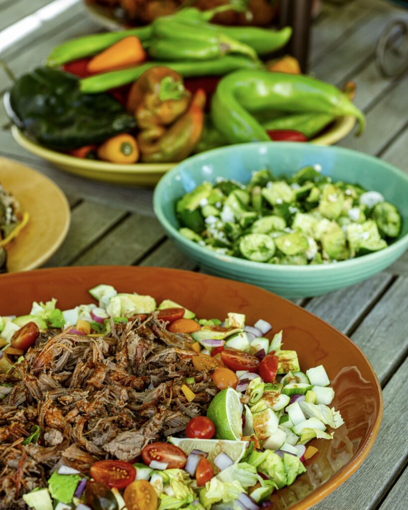 Plates of food on a picnic table with salad in bowls and peppers in the background.