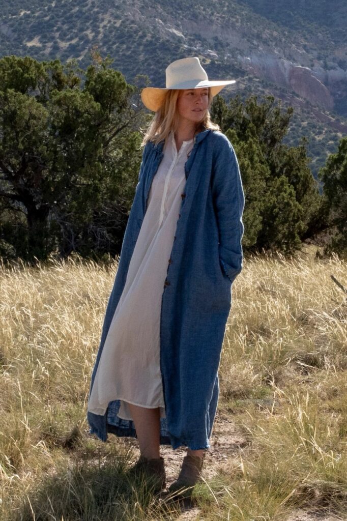 A woman in a long blue coat, tan hat, and long light blue dress in the desert. 