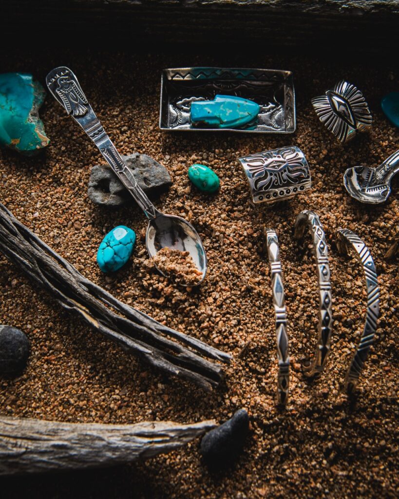 Silver and turquoise jewelry and cutlery in sand.