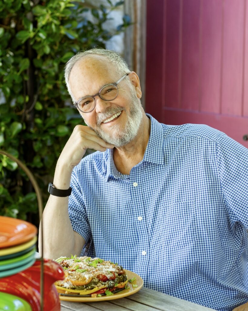 A man sits at a table smiling in a blue button-up and black glasses.
