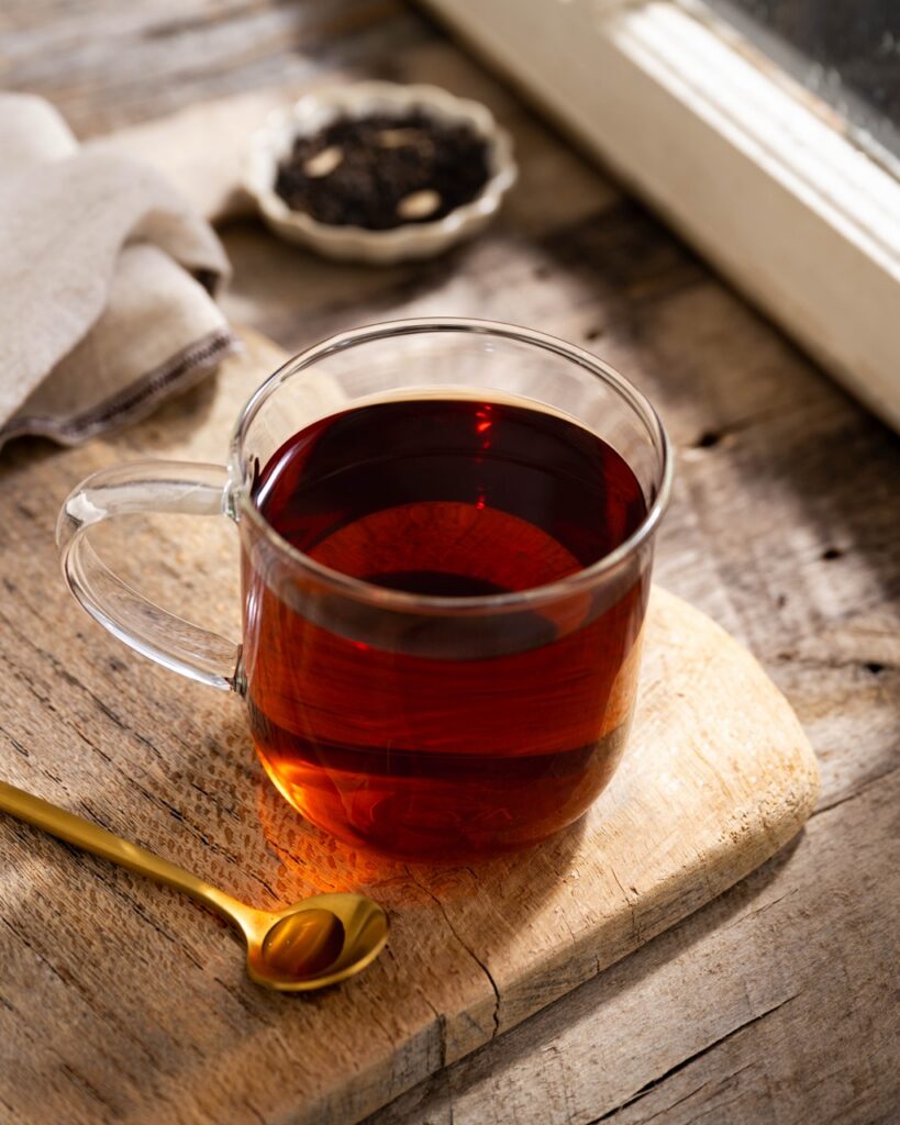 A glass mug of tea on a brown cutting board beside a golden spoon.