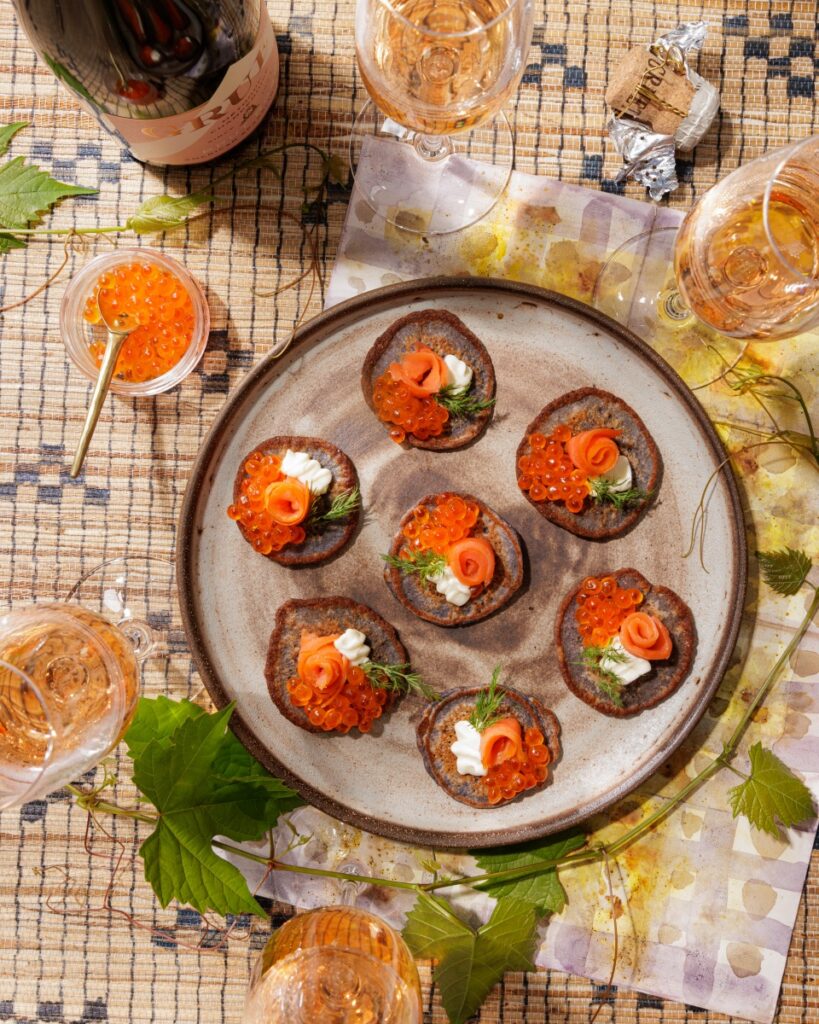 Blue corn blinis topped with crème fraîche, smoked salmon roses, salmon roe, and fresh dill sprigs, served alongside glasses of Gruet Brut Rosé sparkling wine.
