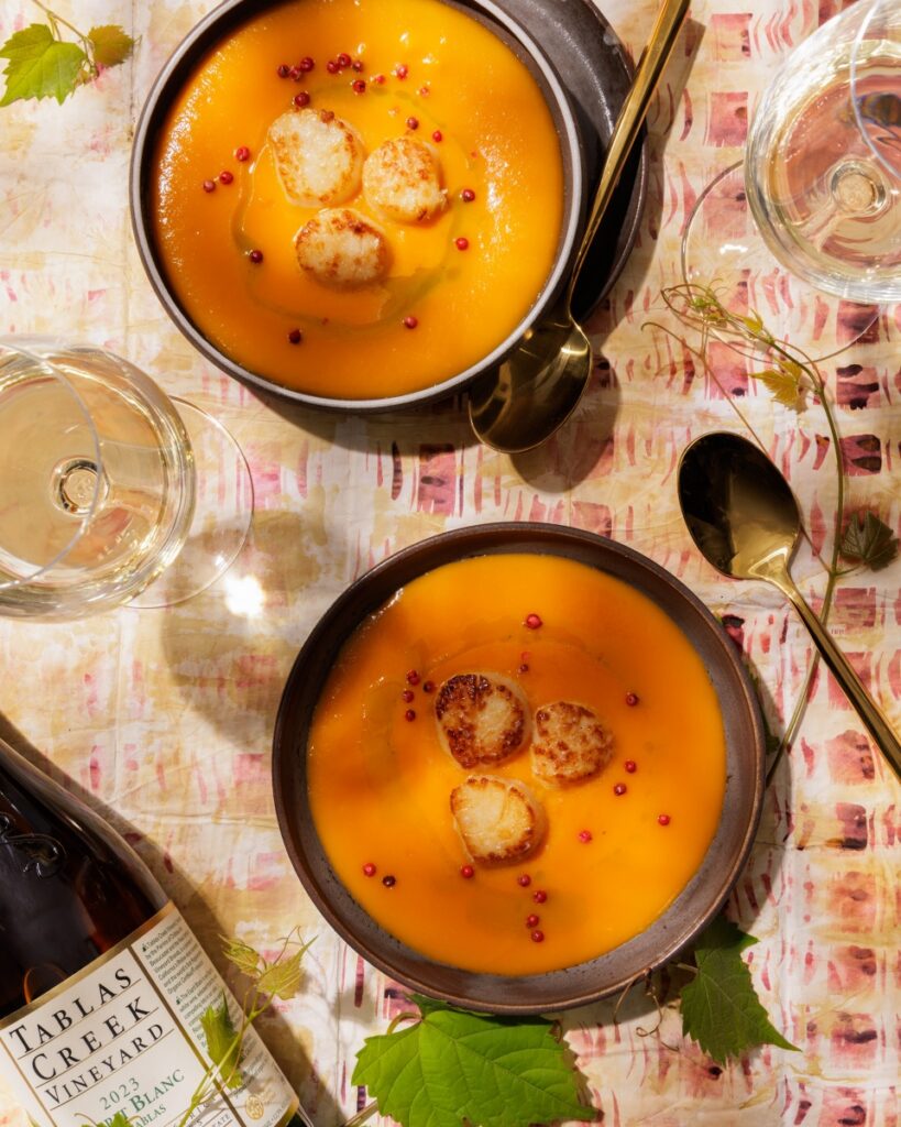 Creamy orange carrot and ginger soup topped with golden-seared scallops, chive oil, and pink peppercorns, served in dark bowls alongside glasses of white wine and a bottle of Tablas Creek Esprit de Tablas Blanc.