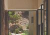 David Naylor Interiors Blends Global Artifacts Into a Sleek Santa Fe Residence Hallway view featuring three hand-carved wooden teak balls sitting on the light-toned stone floor in front of a floor-to-ceiling glass window overlooking a rocky outdoor landscape.