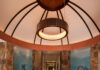 David Naylor Interiors Blends Global Artifacts Into a Sleek Santa Fe Residence Circular dining room under a vaulted ceiling with painted blue and earth-toned wall nichos, a round wooden table, and a dark curved banquette paired with white leather chairs.