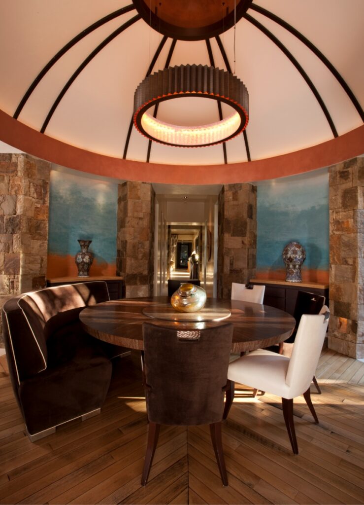 Circular dining room under a vaulted ceiling with painted blue and earth-toned wall nichos, a round wooden table, and a dark curved banquette paired with white leather chairs.
