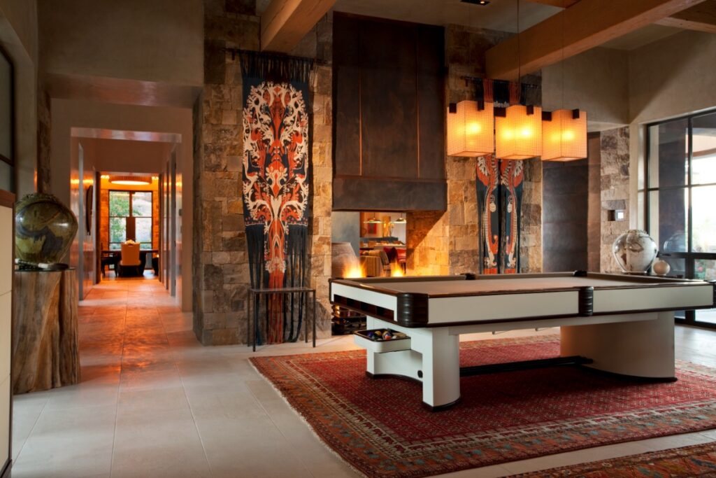 Open living space showcasing a central white-and-dark wood pool table over a red patterned rug, positioned near a hammered-iron fireplace framed by colorful hanging textile art.