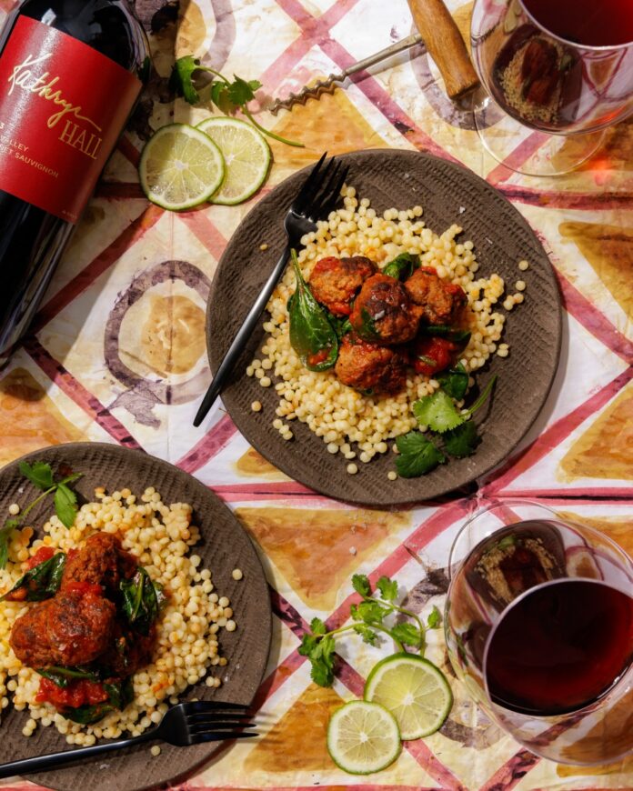 Seared lamb meatballs with wilted spinach simmered in a garlicky tomato sauce, served over giant couscous alongside glasses of red wine and a bottle of Kathryn Hall Cabernet Sauvignon.