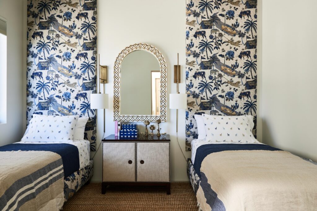 Guest bedroom setup featuring twin beds with blue-and-white Chinoiserie patterned headboards, matching patterned bed bases, neutral throw blankets, a woven floor rug, and a central nightstand below an arched bone-inlay mirror.