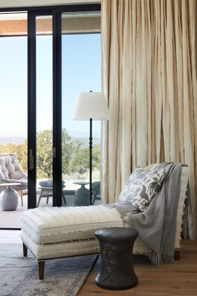 Sitting nook beside floor-to-ceiling glass doors, featuring an upholstered chaise lounge with fringe detailing, a patterned throw pillow, a dark accent stool, and draped cream floor-length curtains.