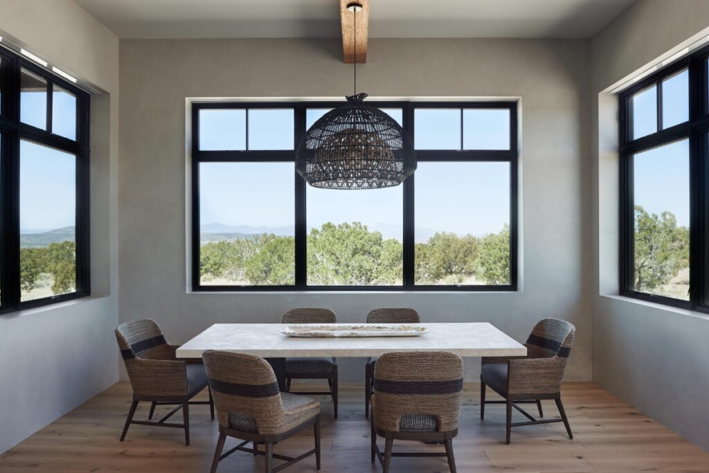 Dining room surrounded by black-framed windows overlooking outdoor hills, furnished with a light stone dining table, woven armchairs, and a dark woven dome pendant fixture hanging from a wood ceiling beam.