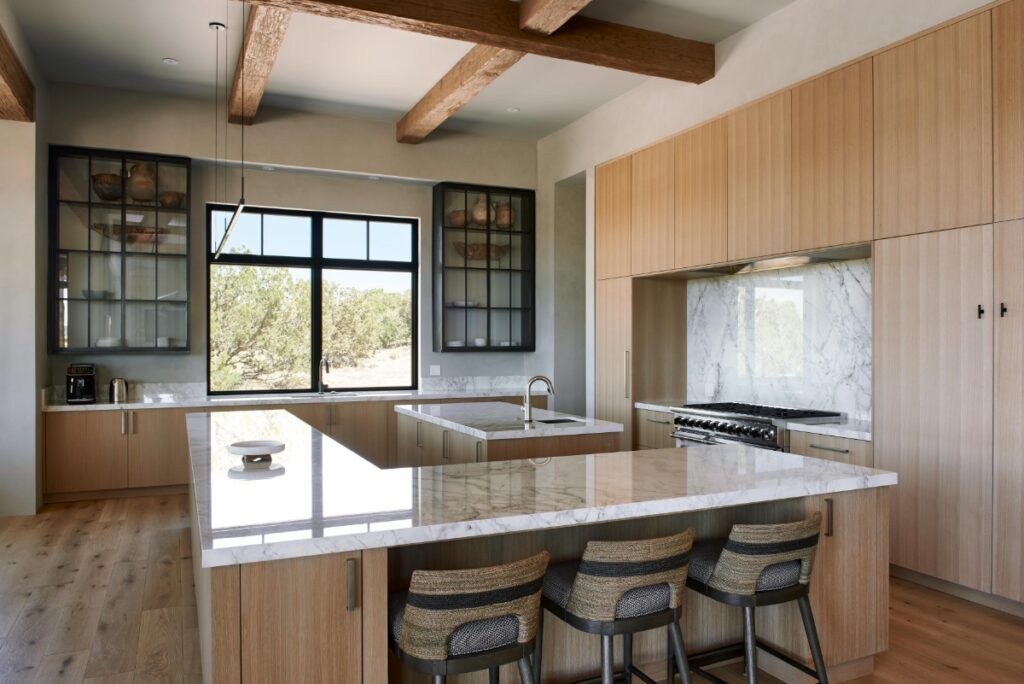 Contemporary kitchen featuring light oak cabinetry, marble countertops and backsplash, a large double island with woven barstools, dark glass-front upper cabinets, and exposed wood ceiling beams.