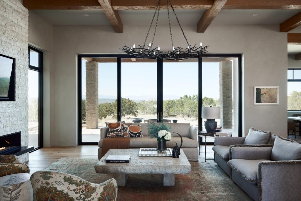 Open-concept living room anchored by a heavy stone coffee table, a branch-style chandelier, neutral sofas, patterned armchairs, exposed ceiling beams, and a white stone fireplace hearth.