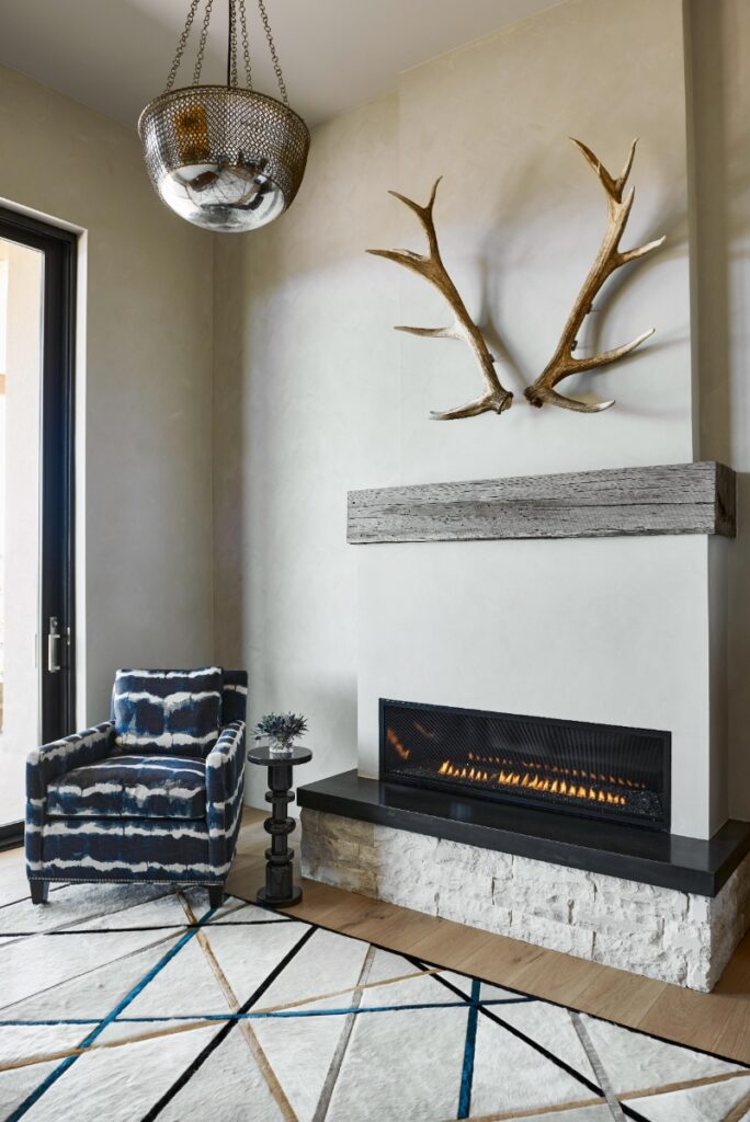 Home study fireplace nook featuring a white lime-wash fireplace with a rustic wooden mantel, a large mounted set of elk antlers above, a modern linear gas fireplace insert, a blue patterned accent chair, and a geometric patterned rug.