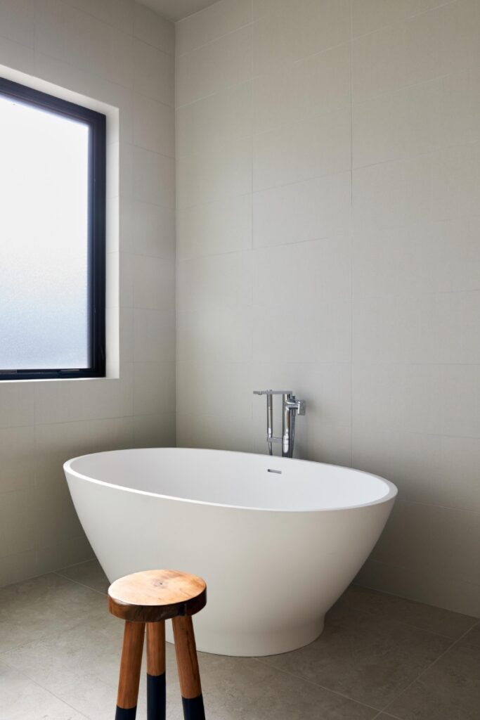 Minimalist bathroom corner featuring a freestanding white oval soaking tub set against smooth neutral tiled walls and floor, paired with a small wooden tripod stool.