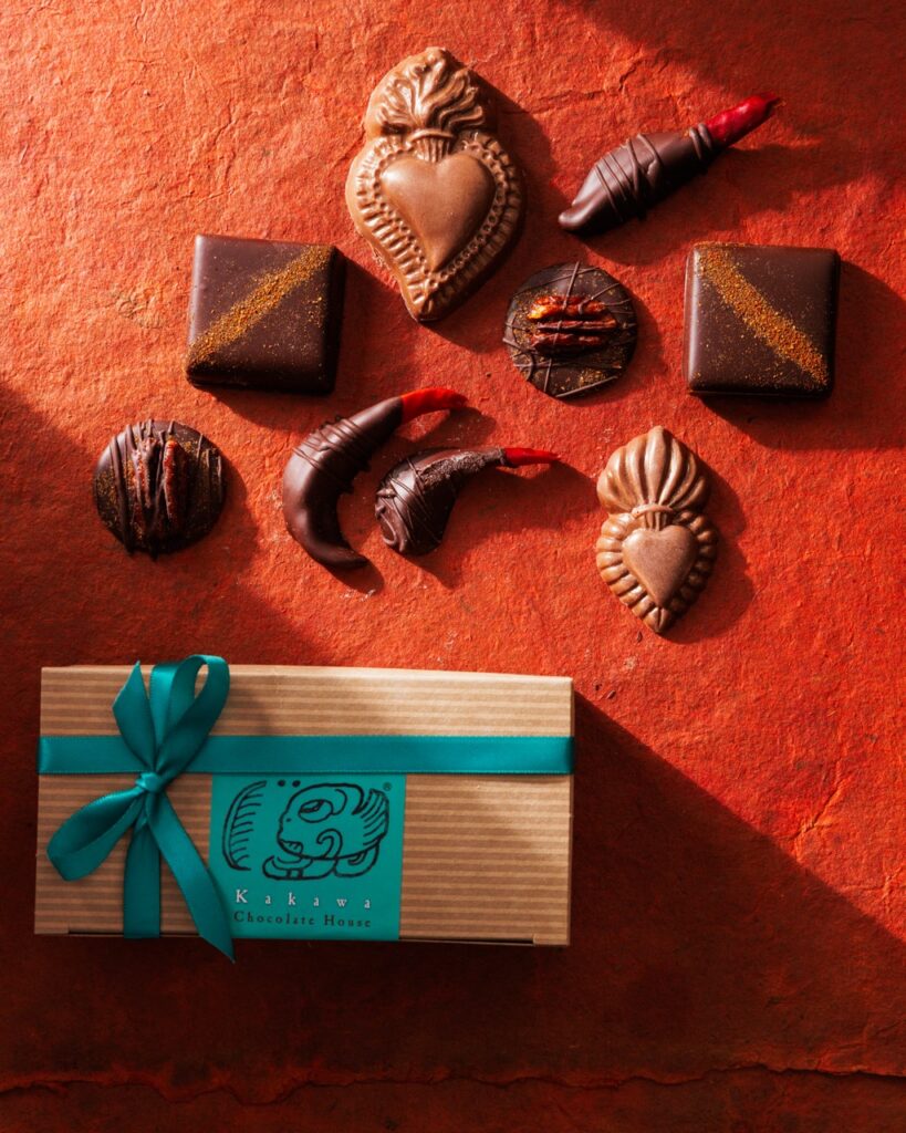 A handful of various shaped chocolates over top of a small brown box of chocolates with a blue ribbon.