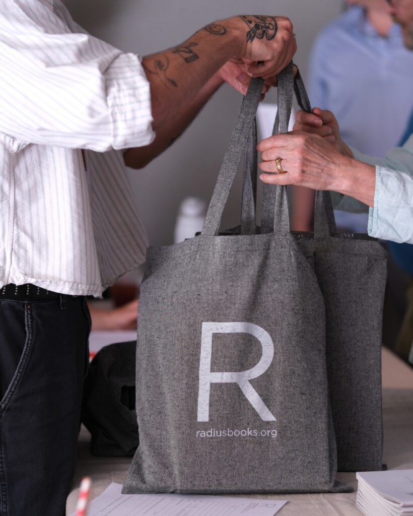 Grey cloth tote bag printed with the white Radius Books logo being handed out at an event.